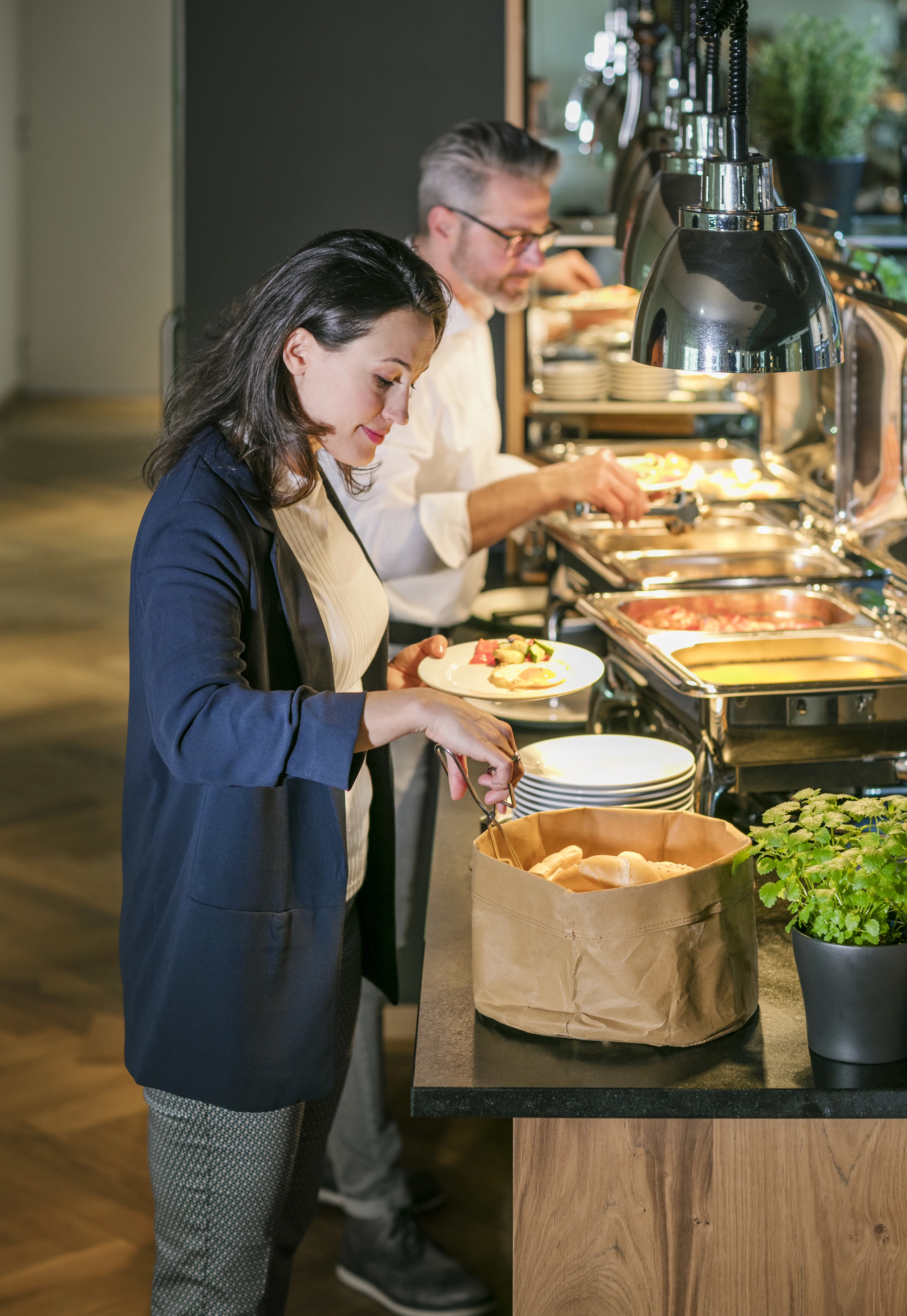 Eine Frau nimmt sich Brötchen an einem Buffet, während ein Mann im Hintergrund serviert.