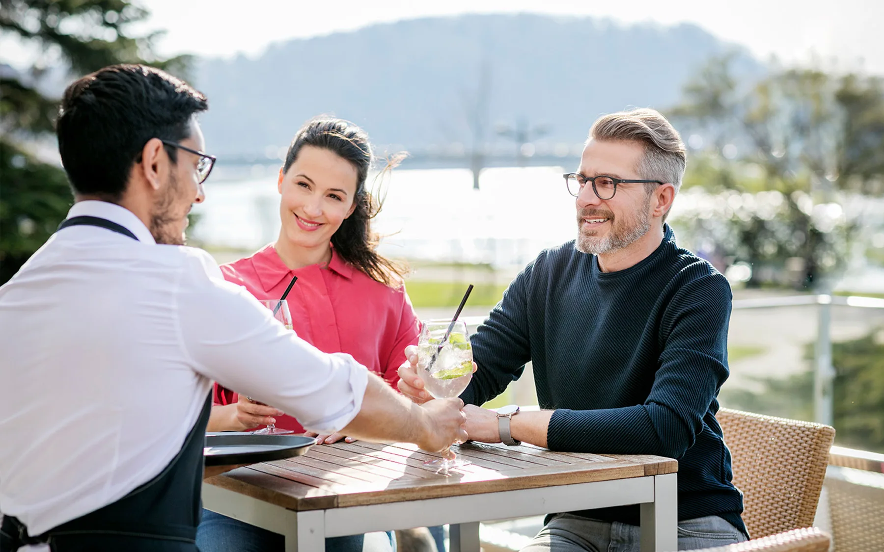 A couple sits at an outdoor table enjoying drinks while a waiter serves them.